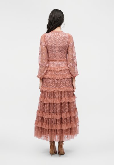 Γυναίκa με μακριά σκούρα μαλλιά φοράει ένα διάφανο μακρυμάνικο ροζ δαντελένιο φόρεμα με στρώσεις και κεντήματα με λουλούδια, και ψηλοτάκουνες σανδάλες, γυρισμένη πλάτη.