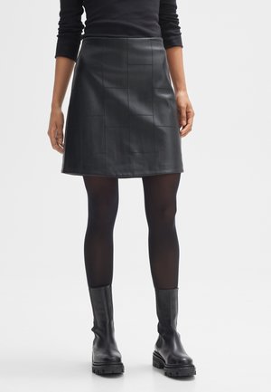 Femme portant un haut noir à manches longues, une jupe en cuir noire avec patchs, des collants noirs et des bottines noires épaisses, debout devant un fond blanc.