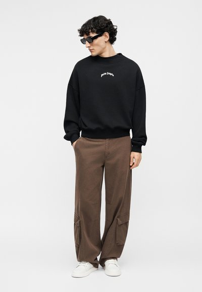 Άνδρας που φοράει μαύρο φούτερ Palm Angels, καφέ παντελόνι cargo, λευκά αθλητικά παπούτσια και μαύρα γυαλιά ηλίου, στέκεται με τα χέρια στις τσέπες.
