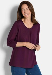 Dunkellila langärmliges Oberteil mit einem runden Ausschnitt und einem gefältelten Frontdetail, kombiniert mit blauen Jeans. Glattes, leichtes Material.
