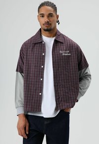 Chemise à carreaux bordeaux et marine avec manches courtes et sous-couche à manches longues grise. Comprend une broderie blanche, une fermeture à boutons et un col.