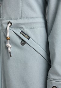 Veste bleu clair avec une texture lisse, poche zippée, cordon tressé et écusson logo. Des boutons-pression viennent compléter le design.