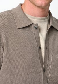 Brun ribbad stickad skjorta med krage, med två svarta knappar och en ficka. Lagd över en ljusbeige crewneck-tröja.