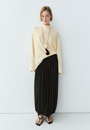Donna che indossa un maglione dolcevita color crema, una lunga gonna nera plissettata, sandali infradito neri e una lunga collana con un grande ciondolo dorato.