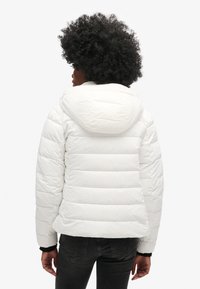 Doudoune blanche avec sections matelassées horizontales, une capuche et des poignets noirs. Texture lisse et isolation légère pour la chaleur.