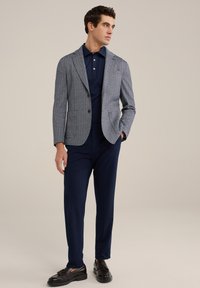 Marineblauwe visgraat blazer over een marineblauwe polo, gecombineerd met donkere broeken en glanzende zwarte instappers, met een slanke pasvorm en soepele texturen.