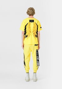 Camiseta gráfica amarilla con estampados de alas en la parte posterior; combinada con joggers amarillos a juego adornados con detalles de etiqueta.