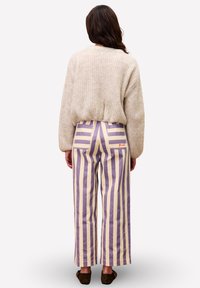 Vrouw in een beige gebreide trui en hoge taille broek met brede pijpen en verticale beige en lavendelstrepen, staand met haar rug naar de kijker gekeerd.