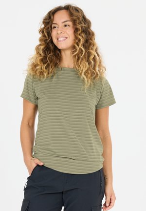 Glimlachende vrouw met krullend haar draagt een groen gestreept shirt met korte mouwen en zwarte broek, staat met één hand in de zak.