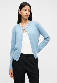 Γυναίκα με γαλάζιο πλεκτό ζακέτα με κουμπιά πάνω από λευκό τοπ, μαύρο παντελόνι, μαργαριταρένιο κολιέ και ασημένια σκουλαρίκια κρίκοι, ποζάροντας με το ένα χέρι στη μέση.