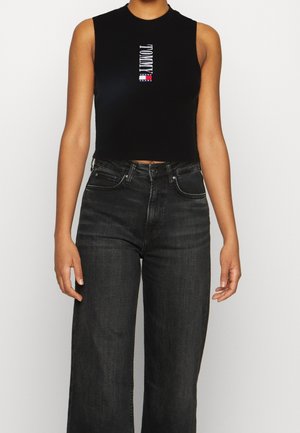 Kobieta ubrana w czarny bezrękawnik typu crop top z pionowym logo „Tommy Jeans” oraz ciemnoszare, wysokowciskowe jeansy o prostych nogawkach.