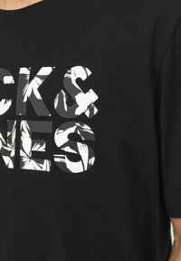 Juodas medvilnės marškinėliai su drąsia grafine spauda, kuri turi baltų gėlių akcentų raidėse "CK & JONES" dideliame blokuotame šrifte.