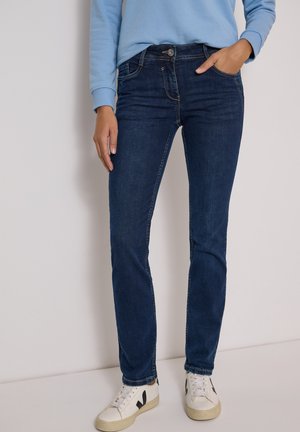 Personne portant un jean slim bleu foncé, un haut à manches longues bleu clair et des baskets blanches, debout devant un fond uni.