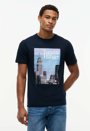Giovane uomo che indossa una t-shirt Tommy Hilfiger blu navy con una grafica dello skyline di New York City e jeans blu, in piedi su uno sfondo bianco.