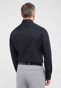 Zwarte longsleeve shirt met een klassieke kraag, een gladde textuur en knoopmanchetten, gedragen met grijze broek en een zwarte riem.