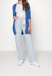 Cardigan largo azul con puños acanalados, usado sobre una camiseta sin mangas blanca. Jeans de talle alto azul claro con un cinturón blanco. Zapatillas blancas.