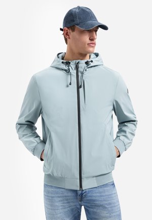 Jonge man in een lichtblauwe capuchonjas met rits en trekkoorden, draagt een donkerblauwe baseballpet, handen in de zakken, zijwaarts kijkend.