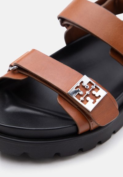 Πέδιλο με μαύρη σόλα και λουριά από ταμπά δέρμα, διακοσμημένο με ασημί μεταλλικό αγκράφα σε σχήμα κομματιού παζλ, σε λευκό φόντο.