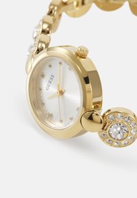 Gold-tone watch with a round face, white textured dial, and crystal hour markers. Features a decorative bracelet link with sparkling accents.