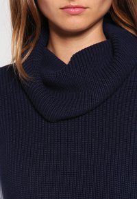 Námořnický pletený svetr s vysokým, přeloženým límcem. Textura je měkká a pletená, s detailním prošíváním po celém povrchu.