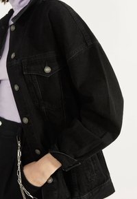 Chaqueta de mezclilla negra con un diseño clásico, botones plateados y un corte relajado con mangas acufadas y bolsillos frontales.