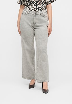 Femme portant un jean gris clair à jambes larges, des talons noirs à bouts pointus et un haut à manches courtes imprimé zèbre, debout devant un fond blanc.