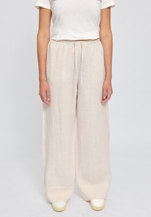 Person wearing loose beige linen pants with elastic waistband and white sneakers, standing against plain light background.