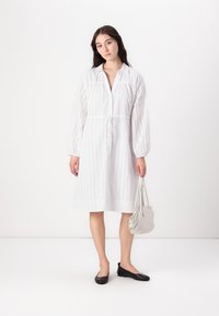 Rochie albă cu detalii din dantelă, croială lejeră, guler, mâneci bufante, talie strânsă, asortată cu o geantă gri deschis și pantofi negri.
