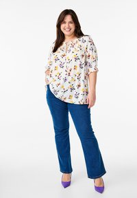 Blusa floreale bianca con motivi multicolori, vestibilità ampia, maniche corte, abbinata a jeans svasati blu scuro e tacchi alti viola.