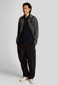 Chaqueta ligera gris sobre una polo negra, combinada con pantalones cargo negros y zapatillas crema. Diseño simple con bolsillos funcionales.