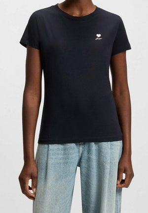 Persoon draagt een zwart T-shirt met korte mouwen met een klein wit hartje en een scriptlogo op de linkerborst, gecombineerd met lichtblauwe jeans.