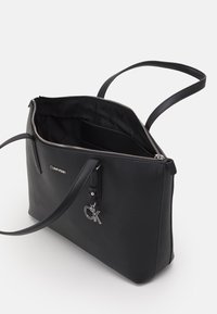 Černá taška typu tote z umělé kůže se stříbrnými kovovými doplňky, horním zipovým uzávěrem a dvěma ramenními popruhy. Uvnitř obsahuje jednu kapsu a podšívku s logem.