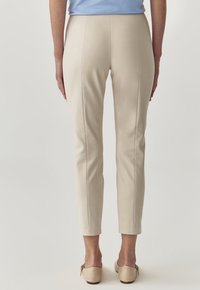 Béžové kalhoty na míru se slim fit střihem, s detaily na předních švech a hladkou texturou. V kombinaci se světle zbarvenými botami.
