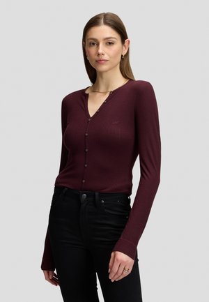 Młoda kobieta z prostymi brązowymi włosami nosi dopasowany bordowy prążkowany top z guzikami i czarne spodnie na jednolitym jasnym tle.