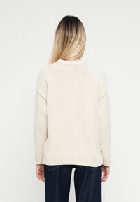 Cremfarbener Strickpullover mit Rundhalsausschnitt und geripptem Saum, mit überschnittenen Schultern und einem lockeren Schnitt, kombiniert mit dunkelblauen Jeans.