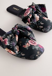 Sandalias tipo slide con base negra, que presentan un gran lazo en la parte superior, tela suave y suela plana.