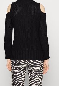 Pull noir à épaules dénudées tricoté, associé à un pantalon imprimé zèbre noir et blanc, vu de dos.