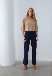 Beige, kurz geschnittenes, geripptes Stricktop mit weiten Ärmeln, kombiniert mit dunkelblauen, hoch taillierten Jeans und beigen Stiefeletten, vor einem neutralen Hintergrund.