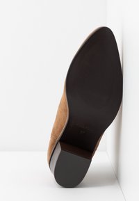 Chaussure habillée en daim marron, présentée en angle pour montrer la semelle en caoutchouc noire avec grip texturé et talon carré, sur fond blanc.
