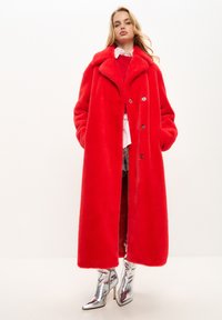 Abrigo largo de piel sintética roja con un amplio cuello y botones frontales, combinado con botas metálicas plateadas hasta el tobillo. Textura suave, silueta oversize.