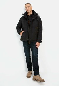 Black insulated jacket with a high collar, multiple pockets, and front zippers. Worn over dark denim jeans and tan boots.