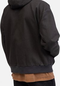 Zwarte hoodie met een relaxed fit, geribde manchetten en onderrand. Textuurstof met een gladde afwerking. Gelaagd over een bruine shirt.