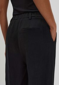Black linen trousers feature a relaxed fit with a flat waistband, adjustable drawstring, and a single rear pocket. Smooth texture, minimalist design.