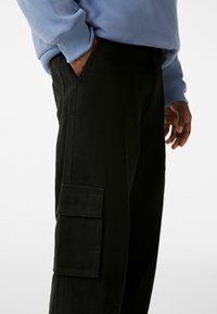Pantaloni cargo neri con tasca laterale, abbinati a una felpa azzurro chiaro. I pantaloni hanno una texture liscia e un design a gamba dritta.