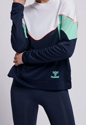 Person iført en sweatshirt med farveblokke i marineblå, hvid og mintgrøn, samt matchende marineblå leggings, stående med armene krydset.