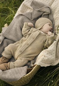 Bež pleteni baby bodi s lesenimi gumbi, v kombinaciji s ujemajočo se kapico. Obdan s teksturiranimi dekicami v mehkobnih tonih, postavljen v pleteno košaro.