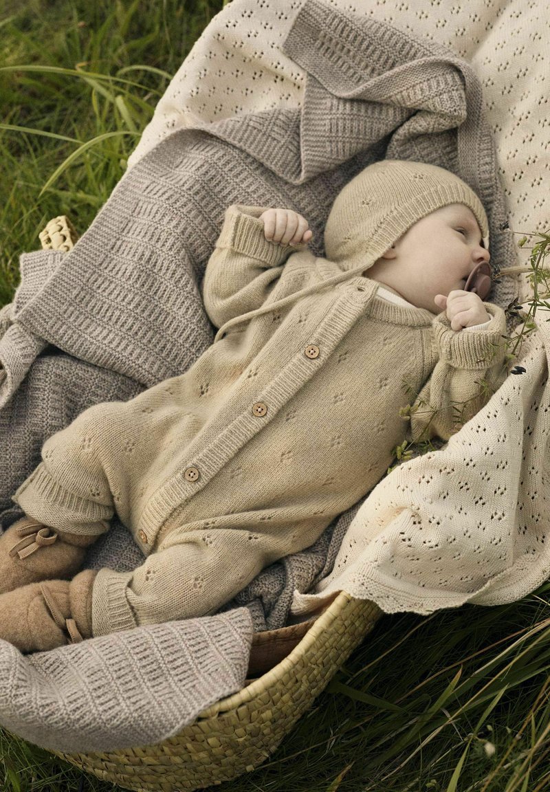 Bež pleteni baby bodi s lesenimi gumbi, v kombinaciji s ujemajočo se kapico. Obdan s teksturiranimi dekicami v mehkobnih tonih, postavljen v pleteno košaro.