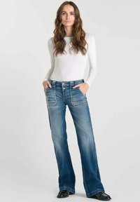 Jeans flare en denim bleu avec un lavage délavé, des coutures détaillées et un design à cinq poches. Associés à une chemise blanche ajustée à manches longues.