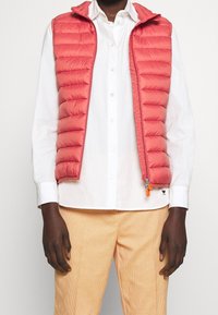 Red puffer vest with horizontal stitching, zip closure, and hood, worn over a white shirt. Paired with light checked pants.
