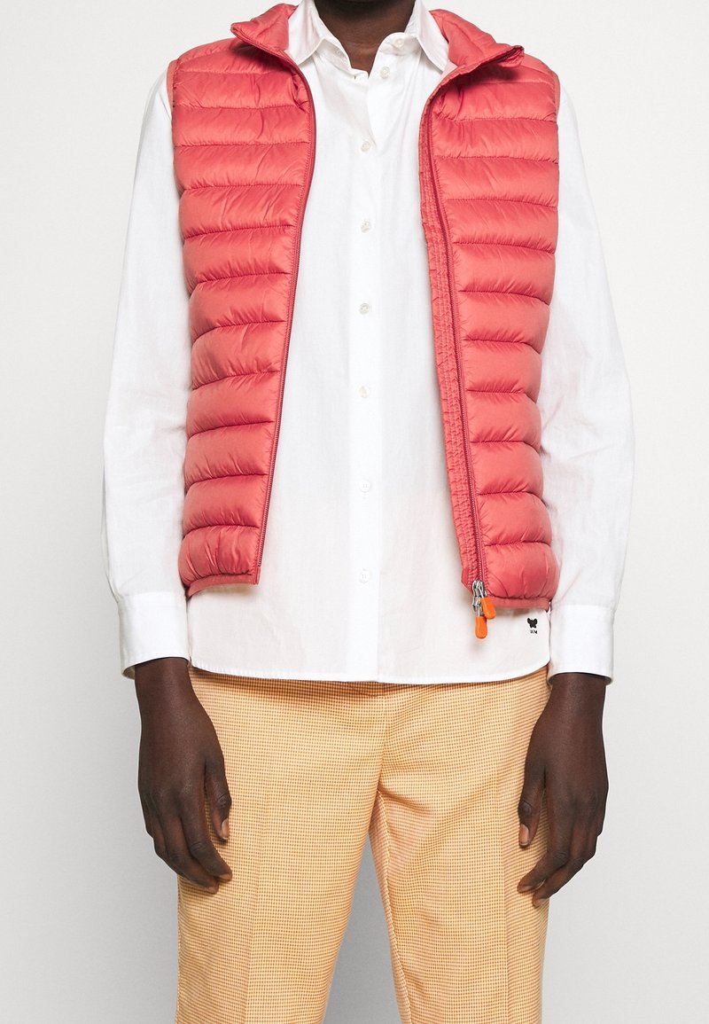 Red puffer vest with horizontal stitching, zip closure, and hood, worn over a white shirt. Paired with light checked pants.